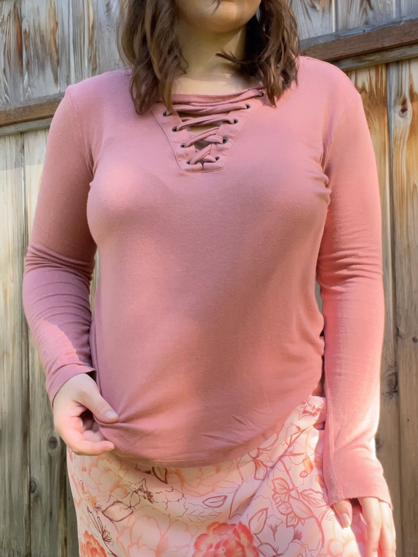 Woman wearing a pink long-sleeve top with lace-up details and a floral skirt standing in front of a wooden fence | Grace Thrifts