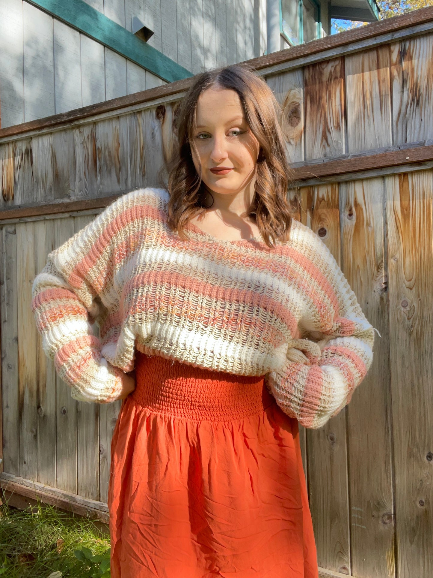 Woman wearing a striped sweater and orange pants standing against a wooden fence | Grace Thrifts