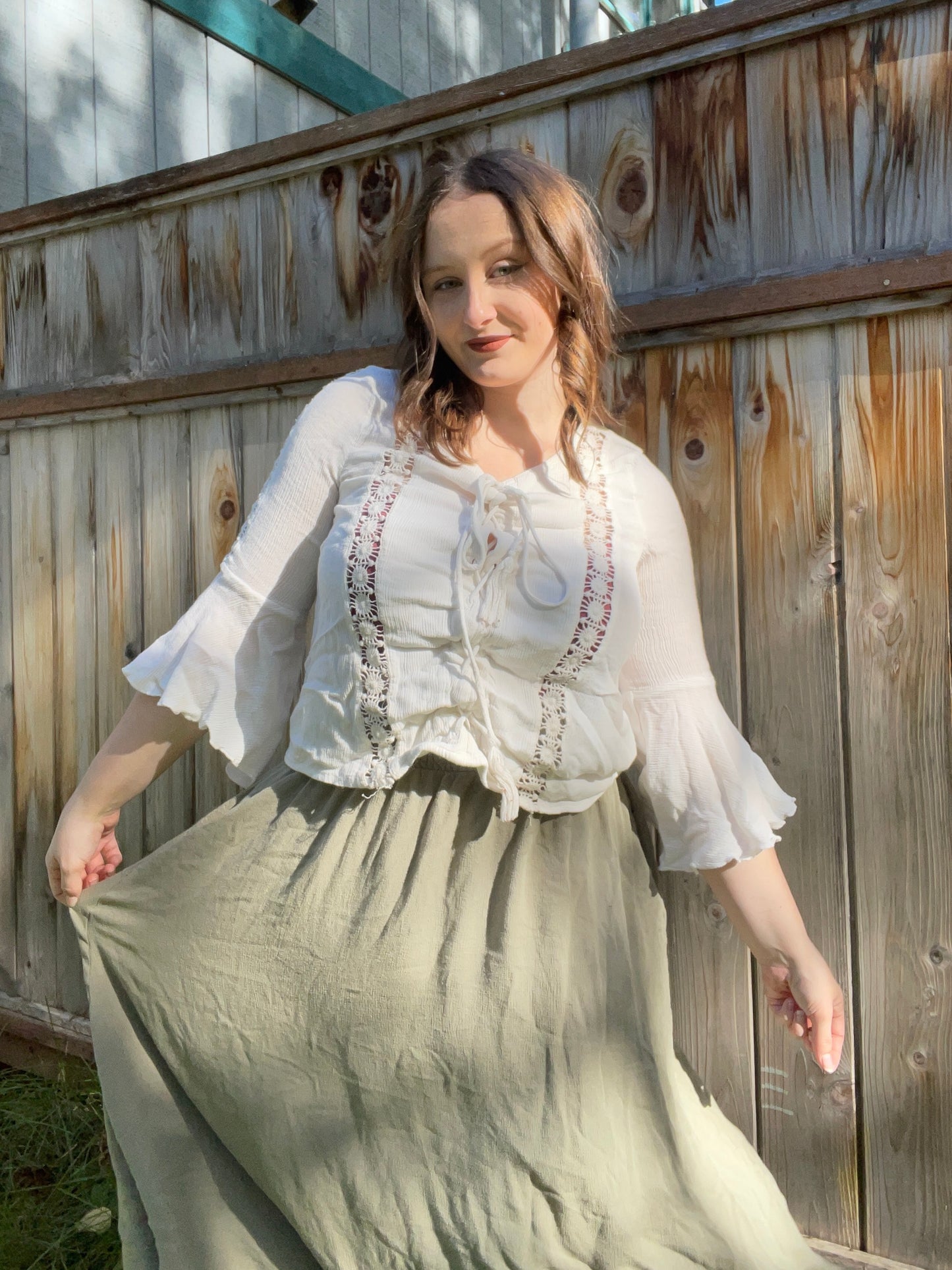 Woman wearing a white blouse with embroidery and a long skirt, standing against a wooden fence.