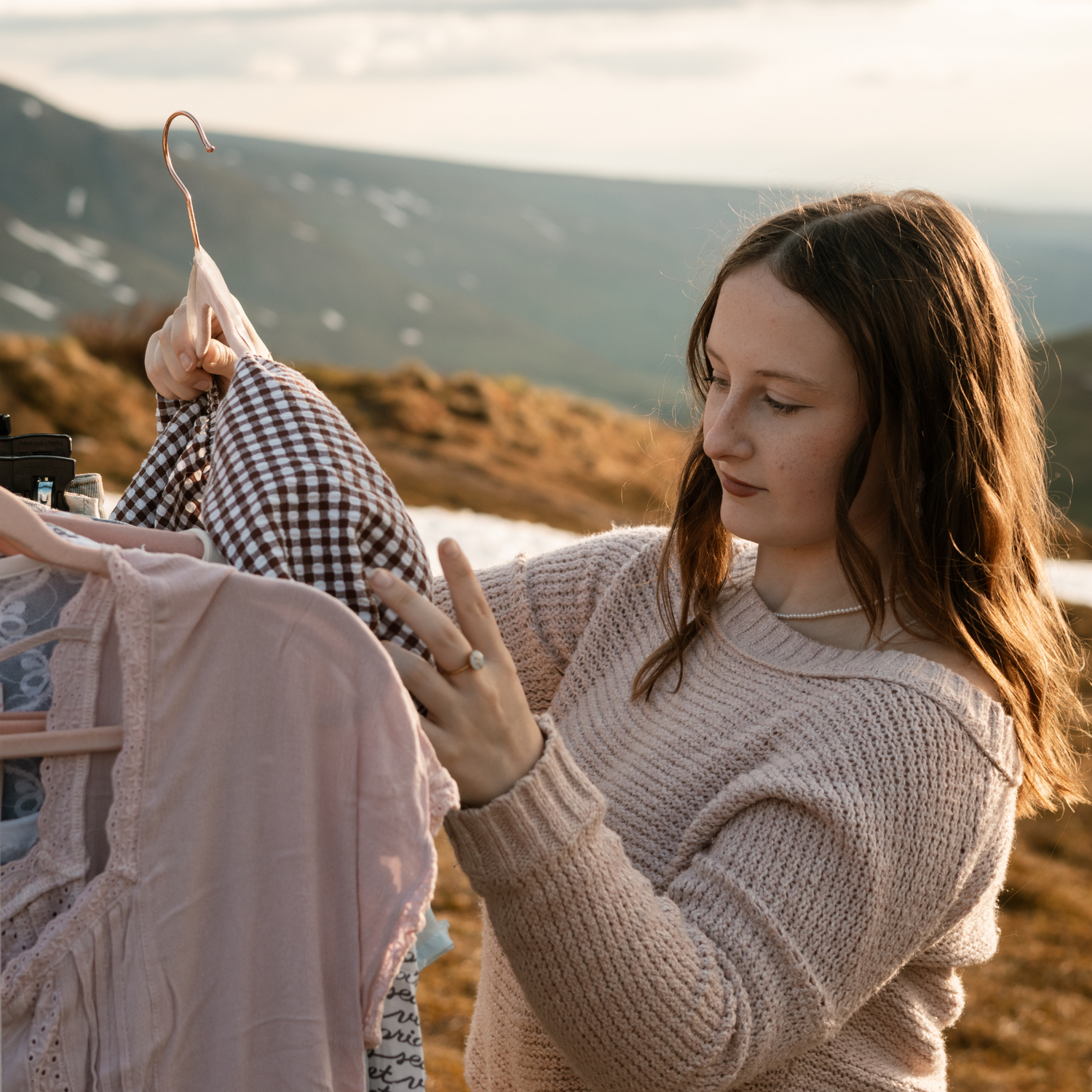 Woman holding a hanger with a checkered shirt against a scenic background. Grace Thrifts