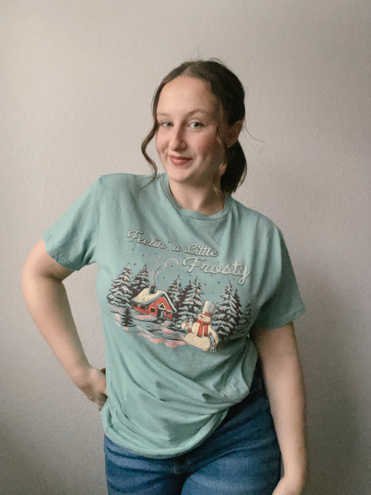 Person wearing a light blue t-shirt with a winter scene design against a plain background Person wearing a green t-shirt and blue jeans against a plain background Feelin' A Little Frosty Graphic Tee | From the Rack | Grace Thrifts