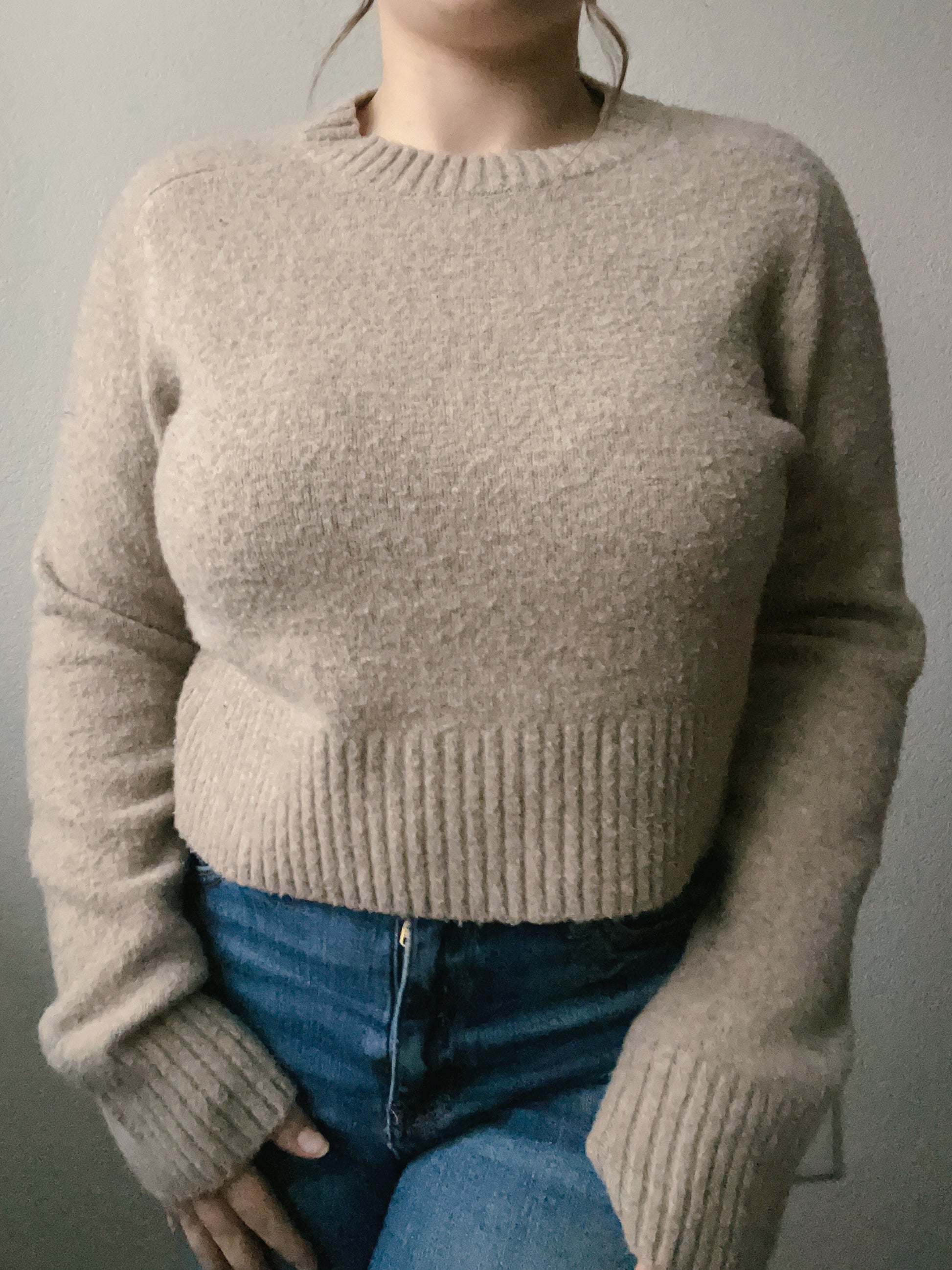 Woman wearing a beige sweater and blue jeans against a plain background Beige Knit Sweater | From the Rack | Grace Thrifts
