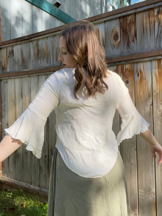 Woman wearing a white blouse with embroidery and a long skirt, standing against a wooden fence.