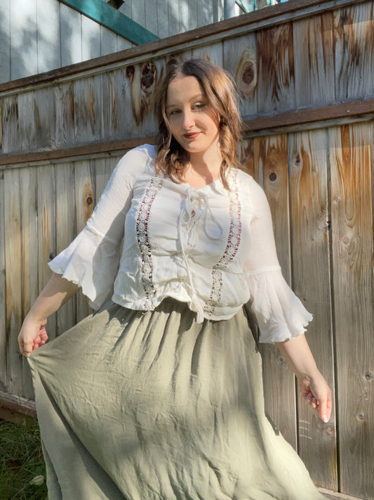 Woman wearing a white blouse with embroidery and a long skirt, standing against a wooden fence.
