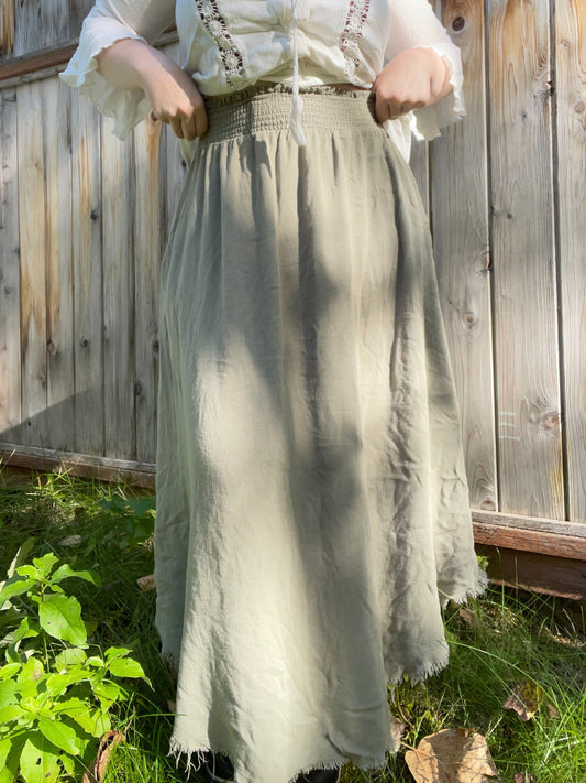 Person wearing a white blouse with embroidery and a long light gray skirt standing in front of a wooden fence.