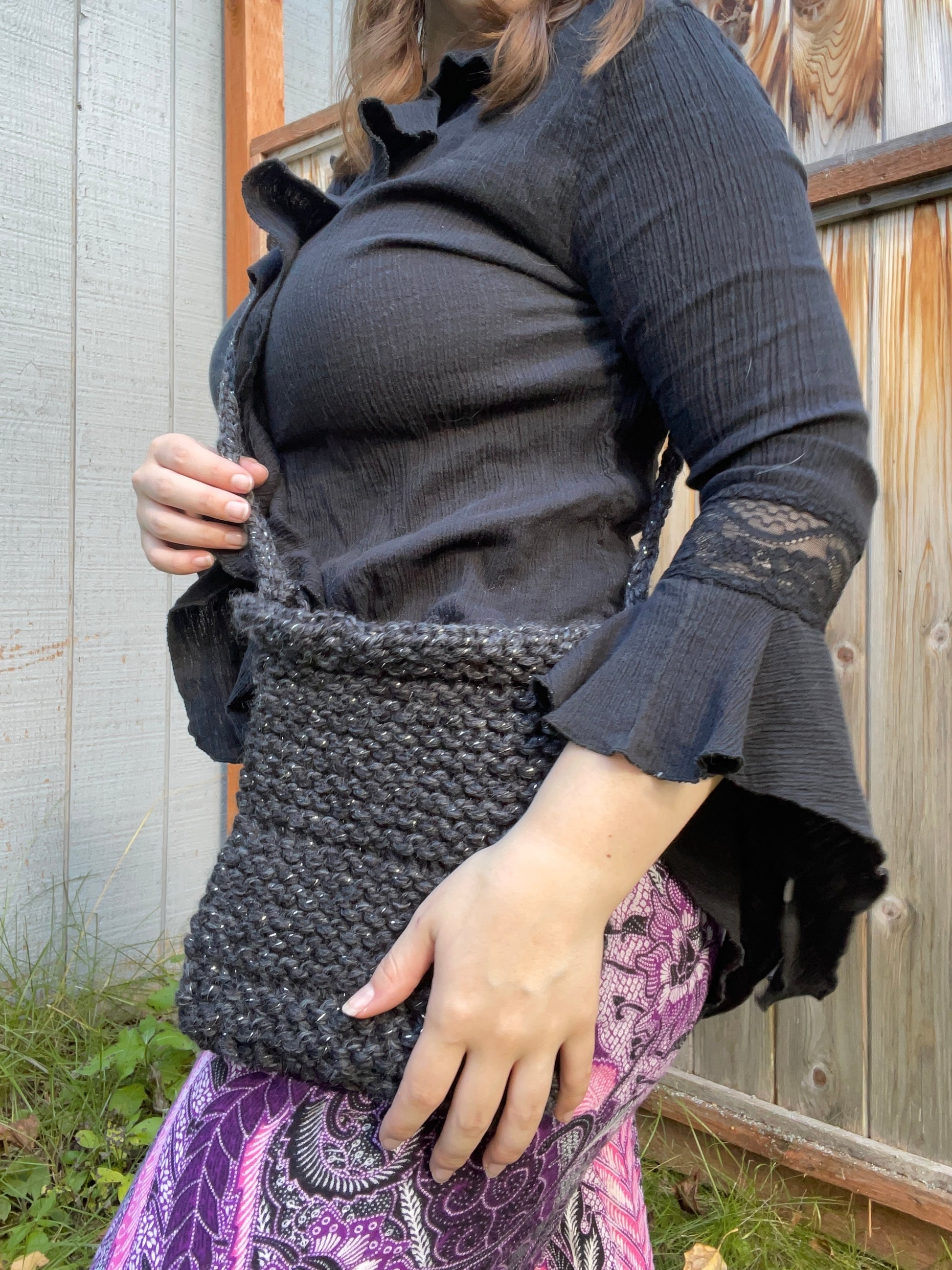Person holding a dark woven bag outdoors against a wooden fence.
