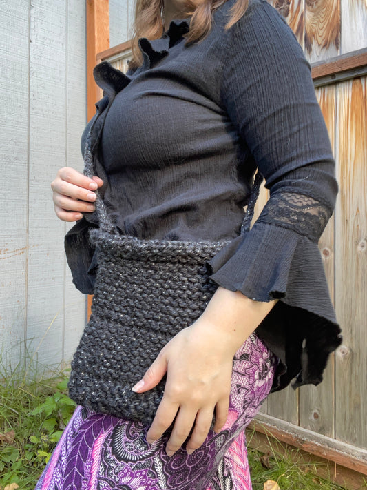 Person holding a dark woven bag outdoors against a wooden fence.
